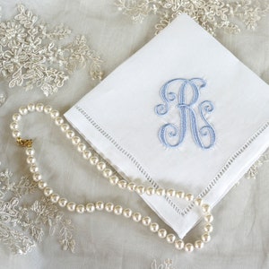 May include: A white linen handkerchief with a light blue embroidered initial "R" on a white lace background with a pearl necklace.