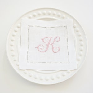 Puede incluir: Una servilleta de lino blanco con una letra "K" bordada en rosa sobre un plato blanco con borde de cuentas.