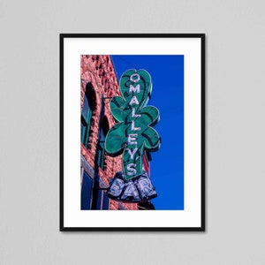 Peut inclure: Une enseigne au néon avec un trèfle vert et les mots "O'Malley's Bar" en lettres blanches. L'enseigne est montée sur un bâtiment en briques avec un ciel bleu en arrière-plan.