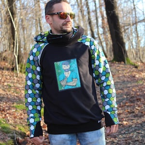 Könnte beinhalten: Schwarzer Kapuzenpullover mit einer Grafik eines tätowierten bärtigen Mannes. Die Ärmel haben ein geometrisches Muster in Grün, Blau und Weiß. Der Hoodie hat einen hohen Kragen und eine schwarze Grundfarbe.
