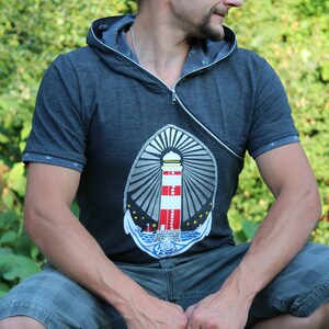 Könnte beinhalten: Dunkelgraues Kapuzenshirt mit Leuchtturm-Grafik. Das Shirt hat einen diagonalen Reißverschluss und kurze Ärmel. Das Leuchtturm-Design zeigt rote, weiße und blaue Farben. Der Text "fusselfreies" ist oben links sichtbar.