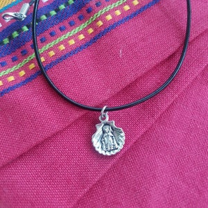 Puede incluir: Collar con colgante de plata que presenta una figura religiosa dentro de una concha de vieira. El colgante cuelga de un fino cord&oacute;n negro con un cierre plateado. El collar descansa sobre una tela roja con rayas bordadas coloridas.
