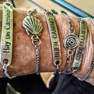 Op de afbeelding: Een verzameling zilverkleurige armbanden met diverse bedels en inscripties. De armbanden hebben rechthoekige labels met zinnen als "Hay Un Camino" en "Buen Camino". Andere bedels zijn een zeeschelp en een spiraalvorm.