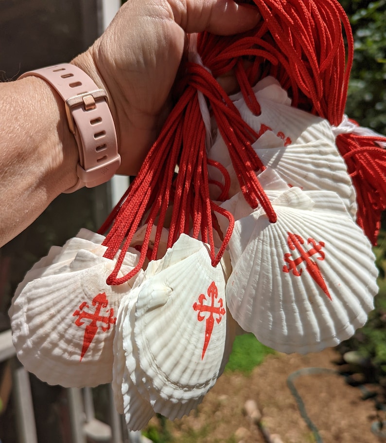 May include: A collection of white scallop shells, each adorned with a red cross emblem, and strung on red cords. The shells are held in a hand, with a pink watch strap visible. These shells are a traditional symbol.