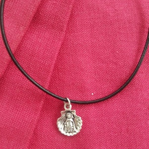 Puede incluir: Collar con colgante plateado que presenta una peque&ntilde;a figura dentro de una concha. El colgante cuelga de un fino cord&oacute;n negro. El collar se encuentra sobre un fondo de tela rosa-rojo vibrante.