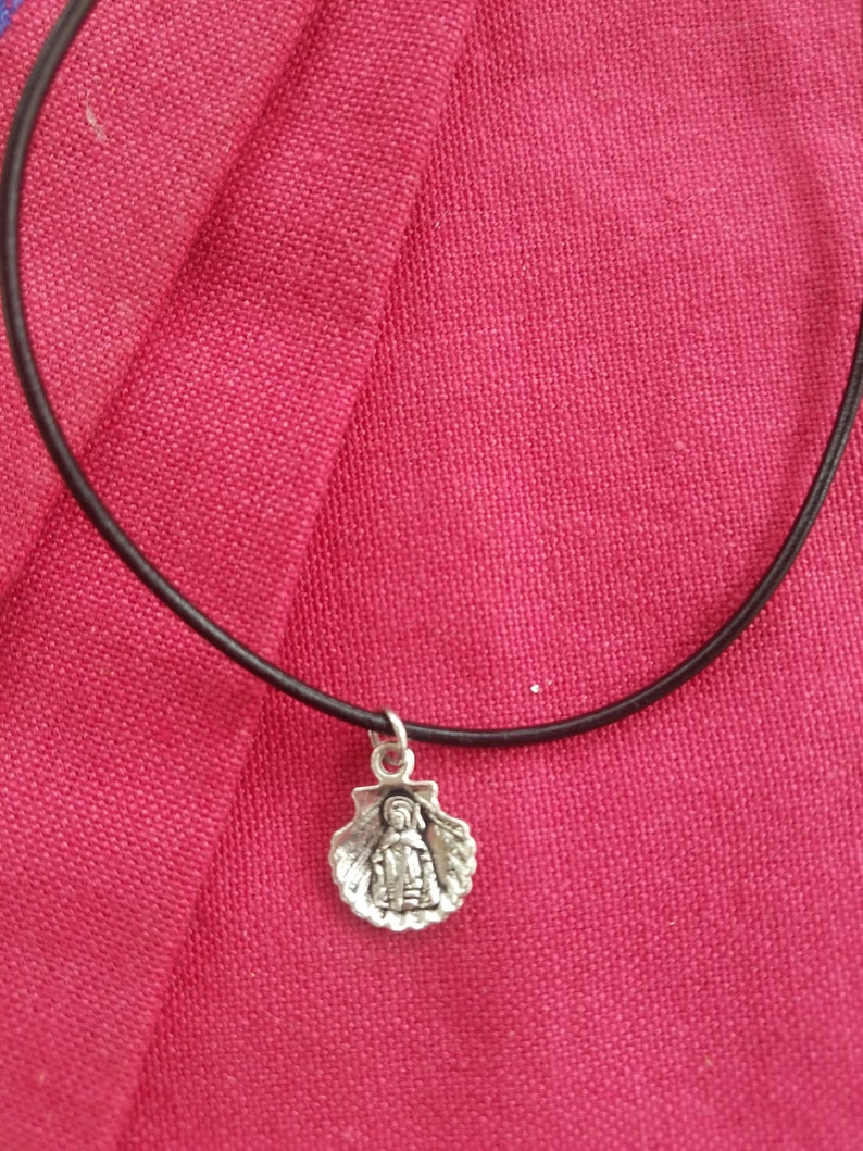 Puede incluir: Collar con colgante plateado que presenta una peque&ntilde;a figura dentro de una concha. El colgante cuelga de un fino cord&oacute;n negro. El collar se encuentra sobre un fondo de tela rosa-rojo vibrante.