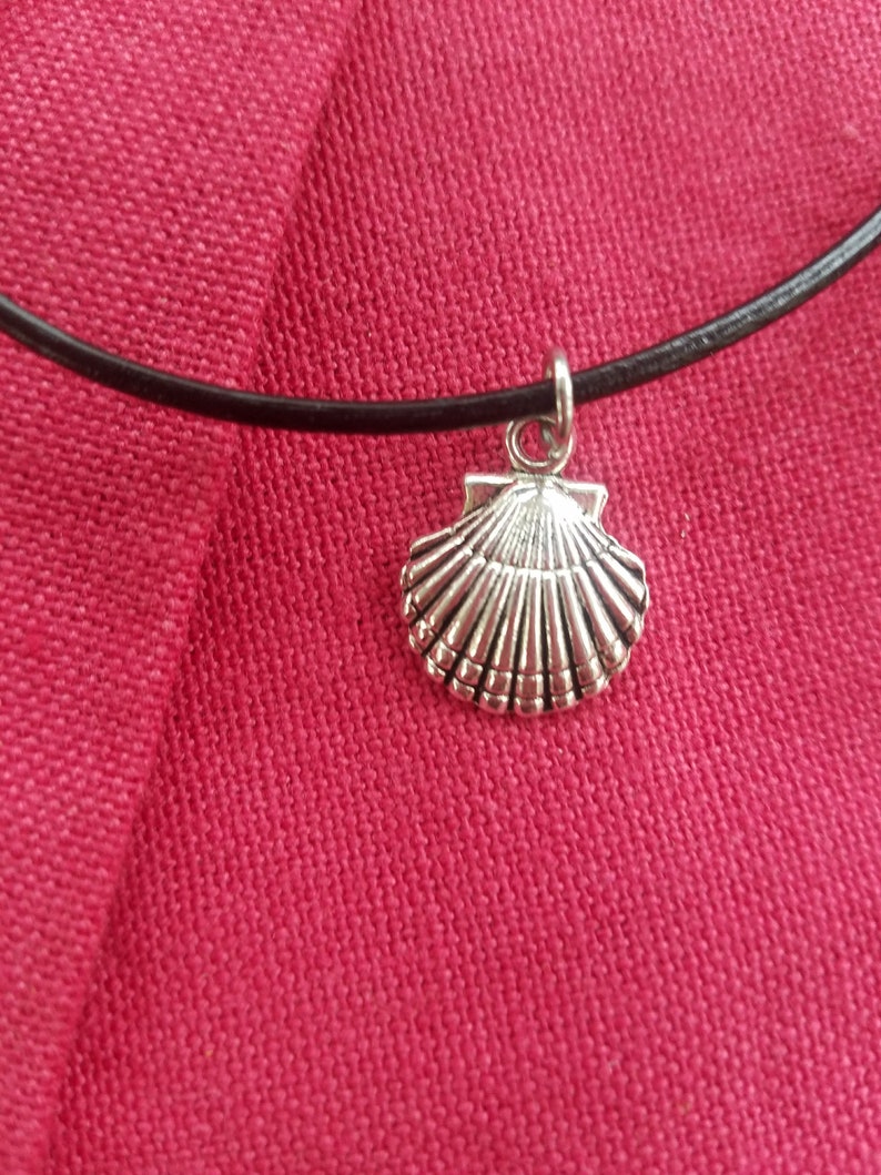 Puede incluir: Un colgante de concha plateada cuelga de un collar de cord&oacute;n negro. El colgante est&aacute; detallado con l&iacute;neas acanaladas y una superficie texturizada. El collar se muestra sobre un fondo rojo vibrante.