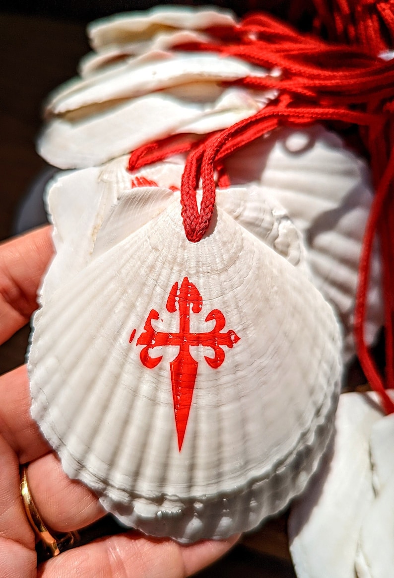 May include: White scallop shells with a red Santiago cross design and red cords. The shells are stacked and held in a hand. The cross is a symbol of the Camino de Santiago pilgrimage.