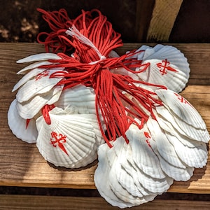 May include: A bundle of white scallop shells, each featuring a red cross emblem, bound with red cord. The shells are displayed on a wooden surface, possibly for a craft or religious purpose.