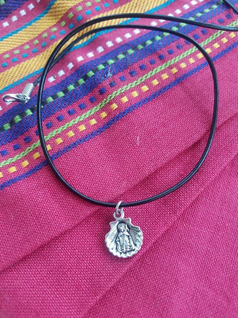 Puede incluir: Collar con colgante de plata que presenta una figura religiosa dentro de una concha de vieira. El colgante cuelga de un fino cord&oacute;n negro con un cierre plateado. El collar descansa sobre una tela roja con rayas bordadas coloridas.