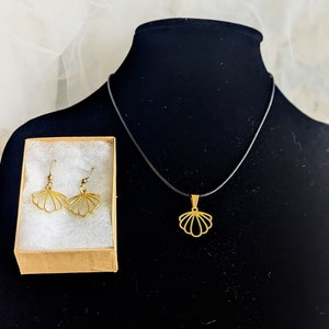 May include: A gold-colored seashell pendant necklace and matching earrings. The earrings are displayed in a small cardboard box with white padding. The necklace has a black cord. The jewelry is displayed on a black mannequin.