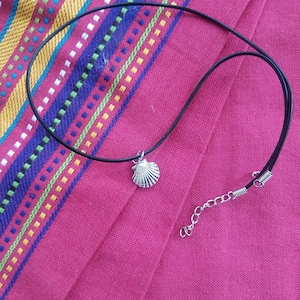 Puede incluir: Collar con colgante de concha plateada y cord&oacute;n negro. El collar se exhibe sobre una tela rosa con rayas coloridas. El collar tiene un cierre de mosquet&oacute;n y una cadena extensora.