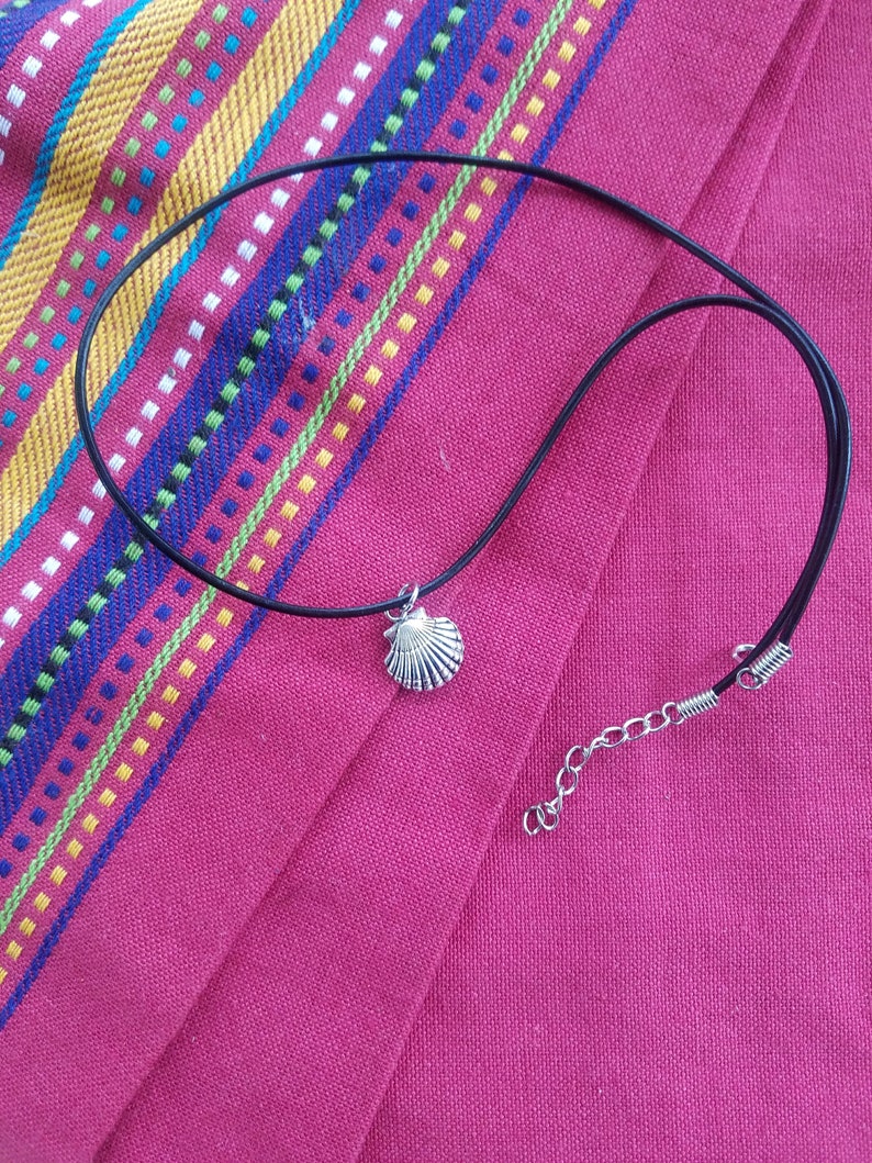 Puede incluir: Collar con colgante de concha plateada y cord&oacute;n negro. El collar se exhibe sobre una tela rosa con rayas coloridas. El collar tiene un cierre de mosquet&oacute;n y una cadena extensora.