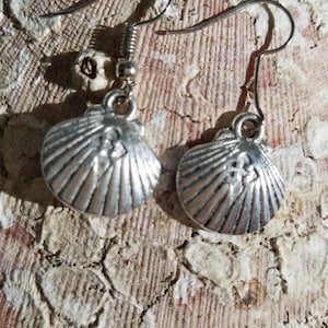 May include: A pair of silver-colored seashell earrings. Each earring features a detailed scallop shell design with a hook for pierced ears. The earrings are displayed on a textured, light brown surface.