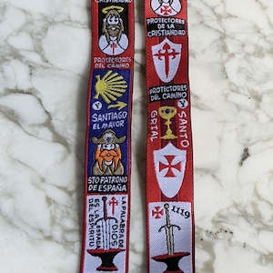 May include: Two red fabric lanyards with various embroidered emblems and text. The lanyards feature religious and historical symbols, including crosses, knights, and the text "Honor Valor Fe".