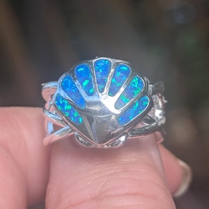 May include: A silver ring featuring a seashell design with a vibrant blue and green opal inlay. The ring has an intricate, interwoven band. The opal's color is reminiscent of the ocean, making it a unique piece of jewelry.