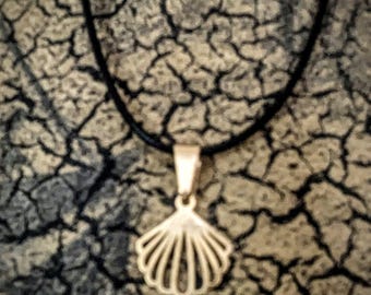 May include: A gold-coloured seashell pendant necklace on a black cord. The pendant is a detailed, openwork design of a fan-shaped shell. The background is a textured, cracked pattern in shades of grey and brown.