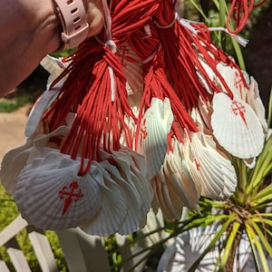 May include: A bundle of white scallop shells, each bearing a red cross symbol, connected by red cords. The shells are held in a hand, with a pink watch strap visible. The background features greenery and a white railing.