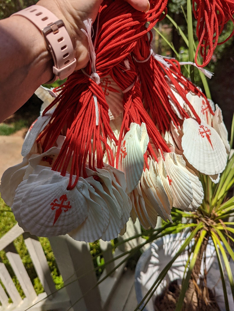 May include: A bundle of white scallop shells, each bearing a red cross symbol, connected by red cords. The shells are held in a hand, with a pink watch strap visible. The background features greenery and a white railing.