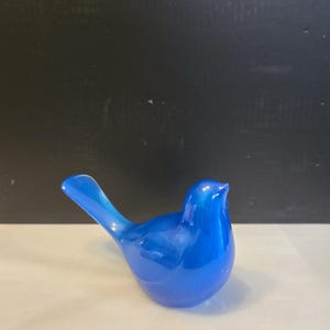 May include: A small, smooth, cobalt blue glass bird figurine. The bird is depicted in a perched position with its head turned slightly upward and its tail feathers angled diagonally toward the upper left. The glass has a translucent, glossy finish.