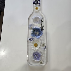 May include: A clear, bottle-shaped resin tray featuring embedded pressed flowers in shades of blue, purple, and white. The design includes a narrow neck with a small hole at the top and a wider base, with various floral arrangements suspended within.