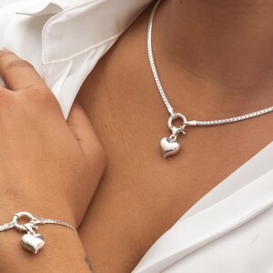 Op de afbeelding: Een zilveren kettingketting en armband met een hartvormige hanger. De ketting en armband zijn beide gemaakt van een delicate, box-kettingstijl.