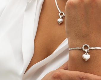 May include: A silver heart pendant necklace and bracelet set. The bracelet is on a wrist, and the necklace is around a neck. Both pieces have a heart-shaped charm. The jewelry is worn over a white shirt.