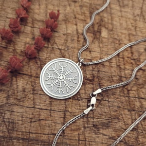 May include: A silver necklace with a circular pendant featuring a Vegvisir symbol. The pendant is engraved with intricate details and the necklace is made of a delicate chain.