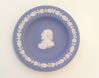 May include: A round, light blue ceramic plate featuring a white relief cameo of a bearded man in profile at the center. The plate is decorated with a white embossed border of oak leaves and acorns encircling the central figure in a circular pattern.