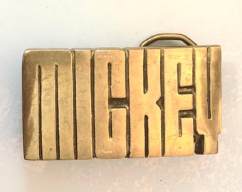 May include: A rectangular brass belt buckle featuring the word MICKEY in large, bold, raised block letters. The metal surface has a polished finish with visible surface scratches and a metal loop attached to the back for belt attachment.