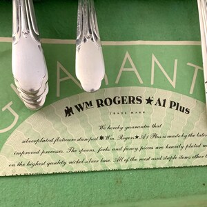 May include: Silver-plated flatware handles resting on a green guarantee card featuring the text WM ROGERS A1 Plus. The card states: We hereby guarantee that silverplated flatware stamped Wm. Rogers A1 Plus is made by the latest and improved processes.