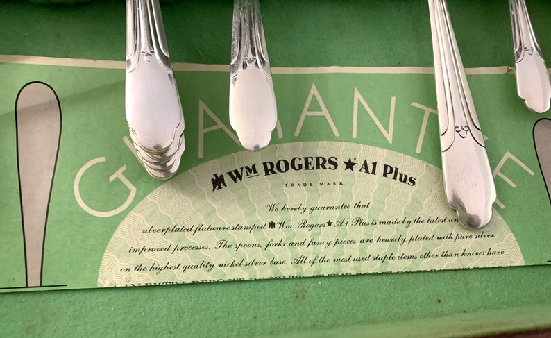 May include: Silver-plated flatware handles resting on a green guarantee card featuring the text WM ROGERS A1 Plus. The card states: We hereby guarantee that silverplated flatware stamped Wm. Rogers A1 Plus is made by the latest and improved processes.