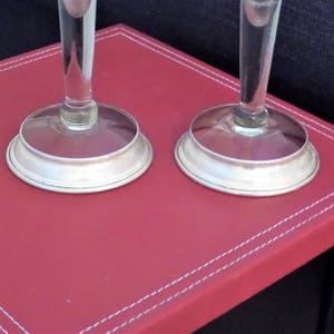 May include: Two clear glass stems with circular silver-toned metal bases. The stems are cylindrical and transparent, connecting to smooth, polished silver-colored circular bases that sit flat. The design is simple, featuring clear glass and metallic silver.