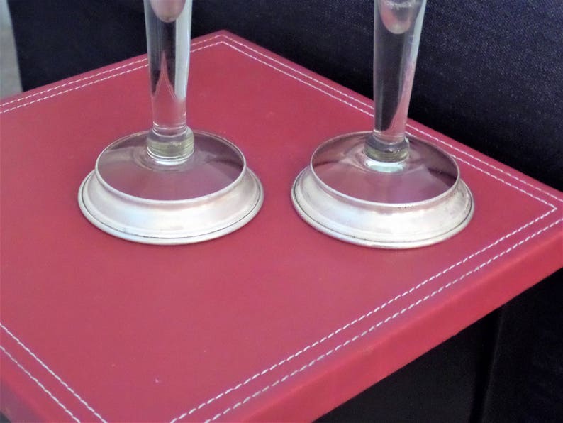 May include: Two clear glass stems with circular silver-toned metal bases. The stems are cylindrical and transparent, connecting to smooth, polished silver-colored circular bases that sit flat. The design is simple, featuring clear glass and metallic silver.