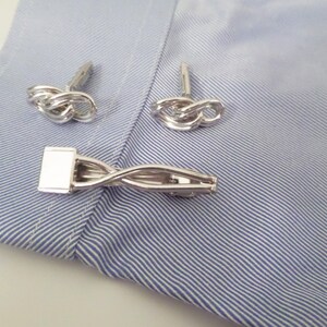 May include: A set of silver-toned accessories consisting of two knot-shaped cufflinks and one tie bar with a twisted metal design. The items are displayed on a light blue and white pinstriped fabric surface, showcasing a polished metallic finish.