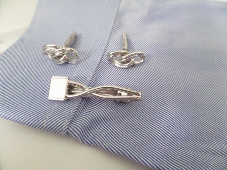 May include: A set of silver-toned accessories consisting of two knot-shaped cufflinks and one tie bar with a twisted metal design. The items are displayed on a light blue and white pinstriped fabric surface, showcasing a polished metallic finish.