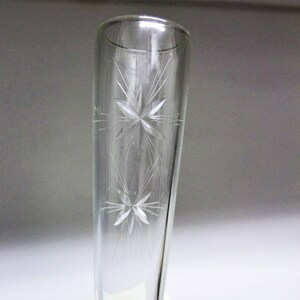 May include: A clear glass bud vase featuring a tapered cylindrical shape and an etched design of two stacked starburst patterns. The glass is transparent and smooth, showcasing the delicate, fine-lined star motifs engraved on the exterior surface.