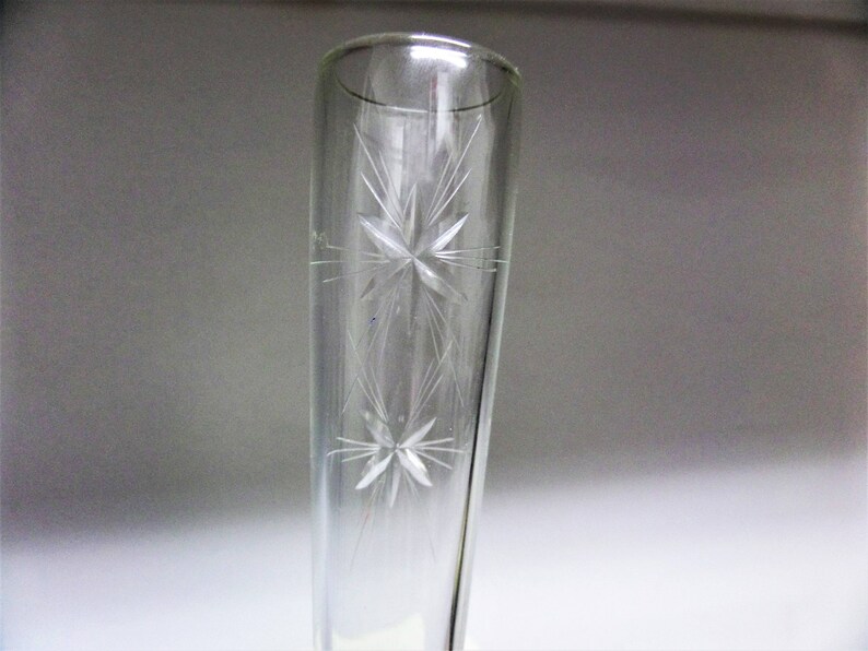 May include: A clear glass bud vase featuring a tapered cylindrical shape and an etched design of two stacked starburst patterns. The glass is transparent and smooth, showcasing the delicate, fine-lined star motifs engraved on the exterior surface.