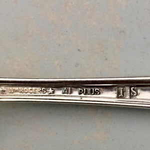 May include: A silver-toned metal utensil handle featuring an engraved hallmark that reads WM ROGERS, a star symbol, A1 PLUS, and the letters I S enclosed in individual square frames. The surface is metallic and reflective with visible text.