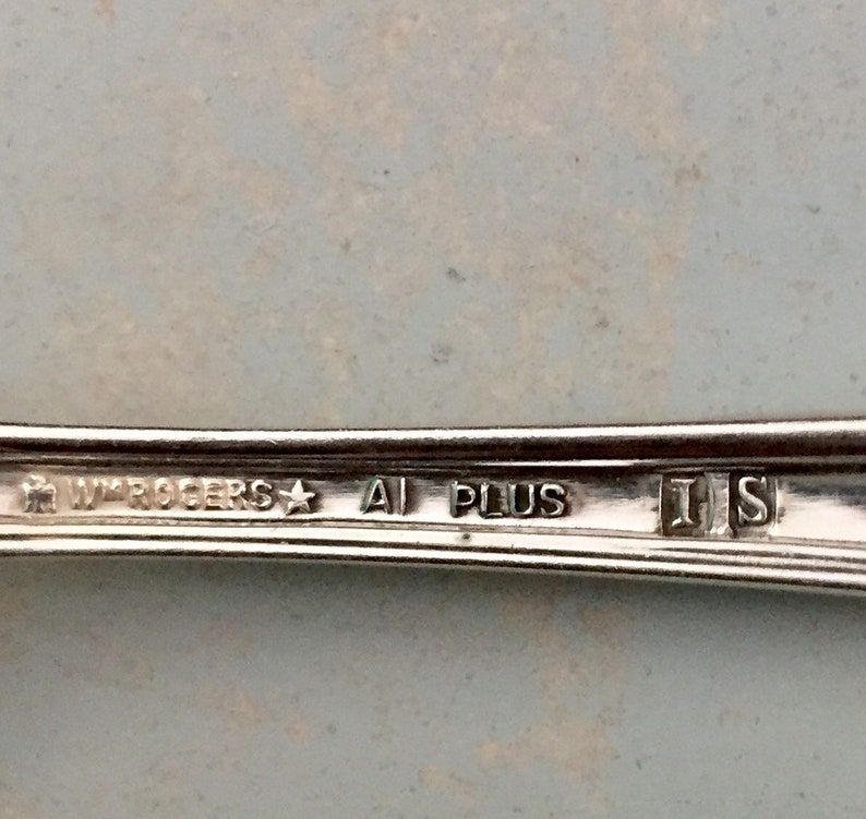May include: A silver-toned metal utensil handle featuring an engraved hallmark that reads WM ROGERS, a star symbol, A1 PLUS, and the letters I S enclosed in individual square frames. The surface is metallic and reflective with visible text.