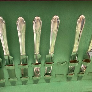 May include: A set of eight silver-toned metal knives with ornate, tapered handles arranged vertically in a storage case lined with light green fabric. Each knife features a smooth, polished blade and a decorative, molded handle design.