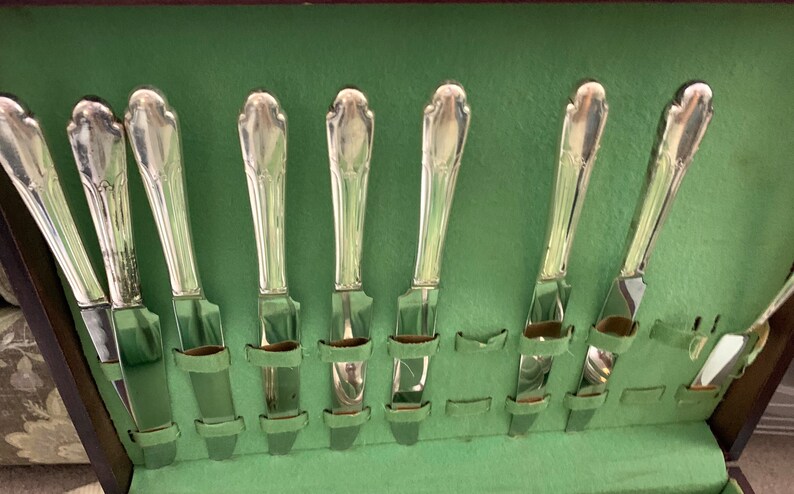 May include: A set of eight silver-toned metal knives with ornate, tapered handles arranged vertically in a storage case lined with light green fabric. Each knife features a smooth, polished blade and a decorative, molded handle design.