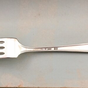 May include: A silver-toned metal fork featuring four tines and a slender handle. The handle is engraved with the text "CHRISTOFLE 20 GRAMS" followed by the initials "HS". The design includes a subtle raised border detail along the handle.