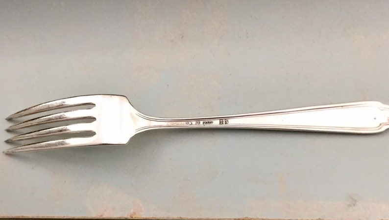 May include: A silver-toned metal fork featuring four tines and a slender handle. The handle is engraved with the text "CHRISTOFLE 20 GRAMS" followed by the initials "HS". The design includes a subtle raised border detail along the handle.