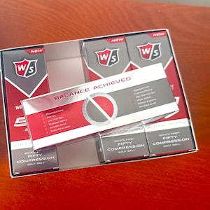 May include: A box of Wilson Staff Fifty Fifty golf balls with red, white, and grey packaging. The box features the text Balance Achieved, Golf's First Fifty Compression Golf Ball, and lists product features including responsive cover and soft feel.