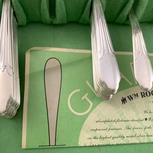 May include: A set of silver-plated flatware handles with a fluted design, arranged in a green felt-lined case. A paper insert is visible with the text WM ROGERS and a graphic of a spoon, detailing the silver-plated flatware and nickel-silver base.