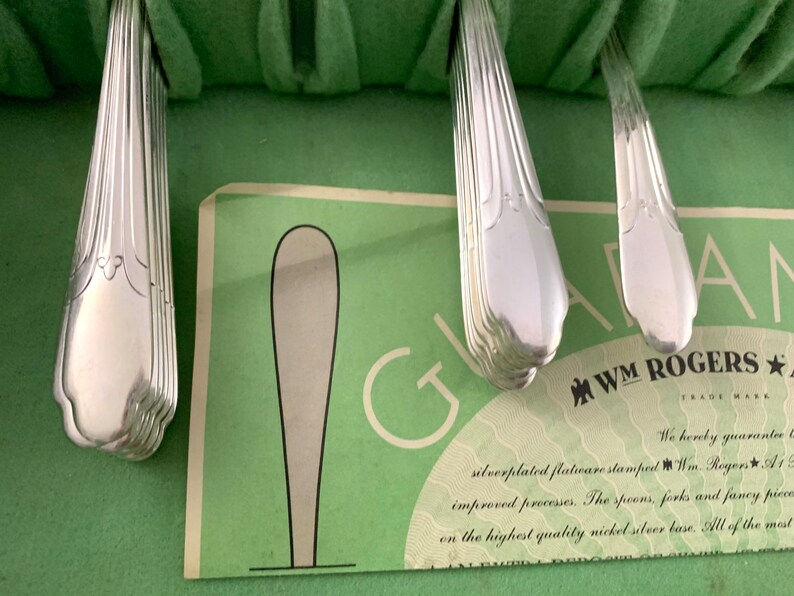 May include: A set of silver-plated flatware handles with a fluted design, arranged in a green felt-lined case. A paper insert is visible with the text WM ROGERS and a graphic of a spoon, detailing the silver-plated flatware and nickel-silver base.