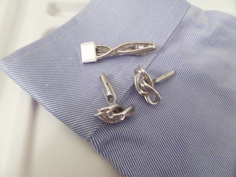 May include: A set of silver-toned metal accessories consisting of a tie bar with a twisted design and two matching cufflinks featuring an intricate knotted loop pattern, all displayed on a light blue and white pinstriped fabric cuff.