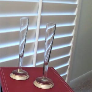 May include: Two tall, slender clear glass bud vases with etched floral details on the upper sections. Each vase features a narrow cylindrical body that flares slightly at the top, supported by a circular, metallic-toned base at the bottom.