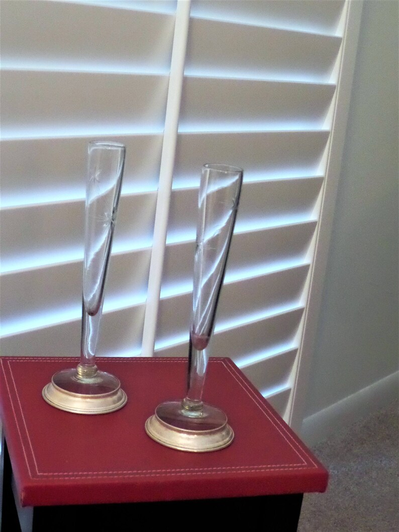 May include: Two tall, slender clear glass bud vases with etched floral details on the upper sections. Each vase features a narrow cylindrical body that flares slightly at the top, supported by a circular, metallic-toned base at the bottom.